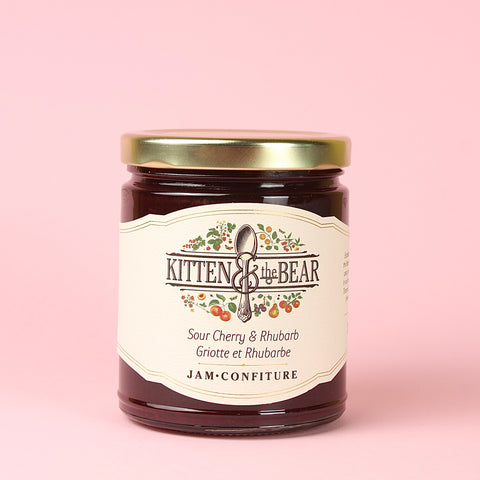 A glass jar of Kitten & the Bear Sour Cherry & Rhubarb jam with a gold metal lid and cream-colored label, set against a solid light pink background.