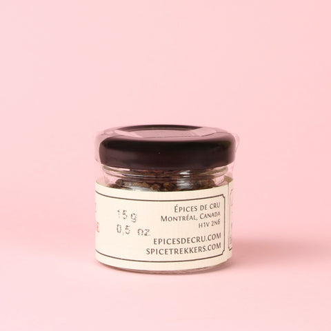 A small clear glass jar of Wild Grains of Paradise with a black lid and a cream-colored label, set against a solid light pink background. The label features black text reading 'ÉPICES DE CRU', 'MONTRÉAL, CANADA', 'HIV 216', 'EPICESDECRU.